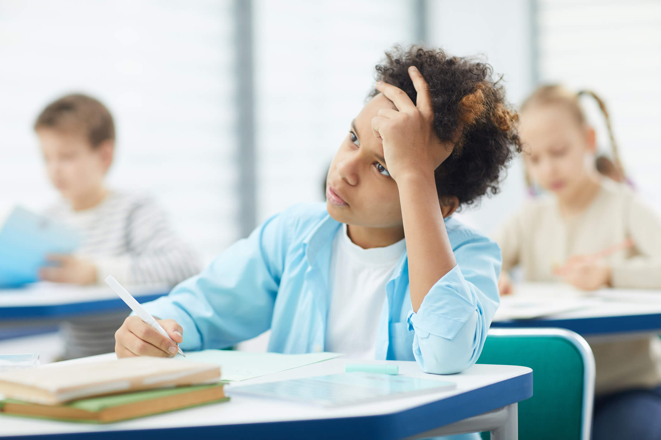 A child in school with dark curly hair and a light blue collared shirt over a white tee looks up while taking notes with one hand on their forehead.