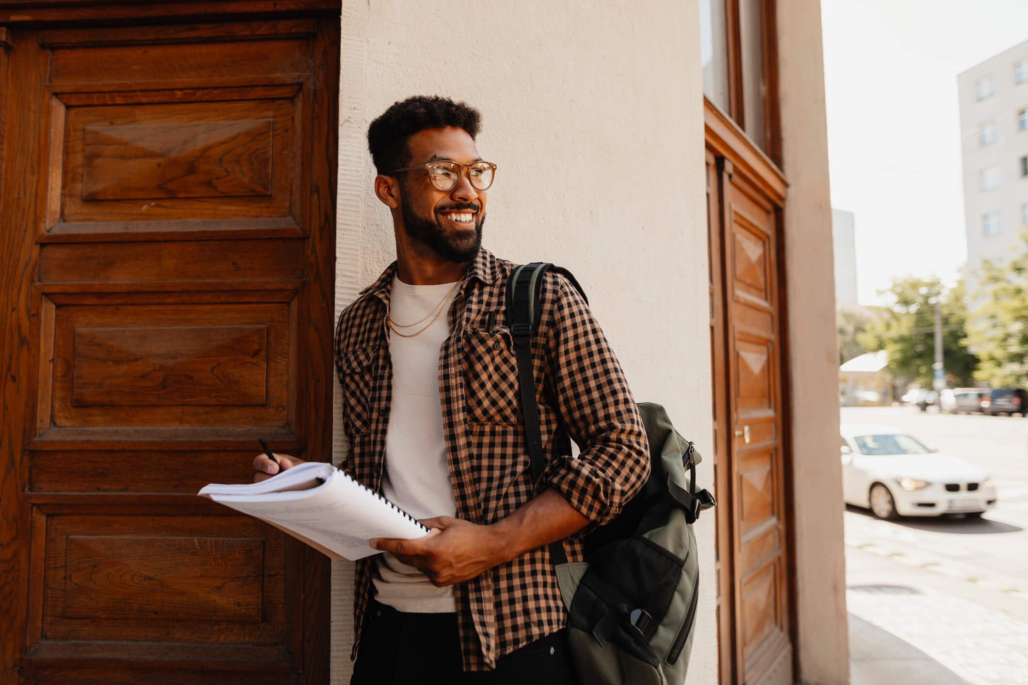 A man in a white shirt and checkered long-sleeve shirt wearing glasses is standing outside a doorway; he is smiling while holding a notebook and looking past him.