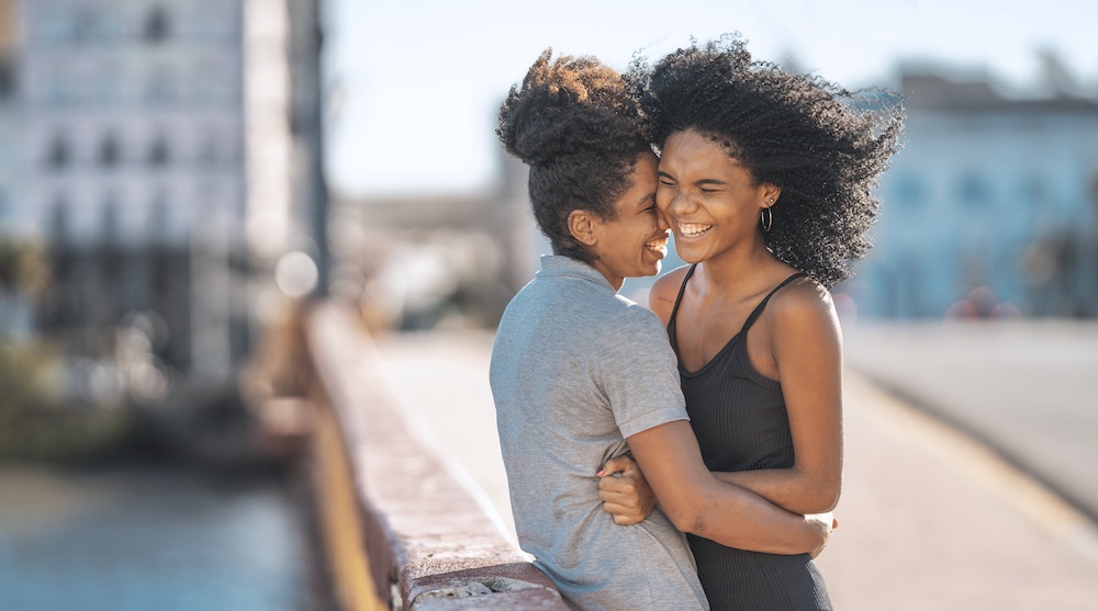 Two women are standing outside on a bridge; they are standing close and embracing, both of them are smiling.