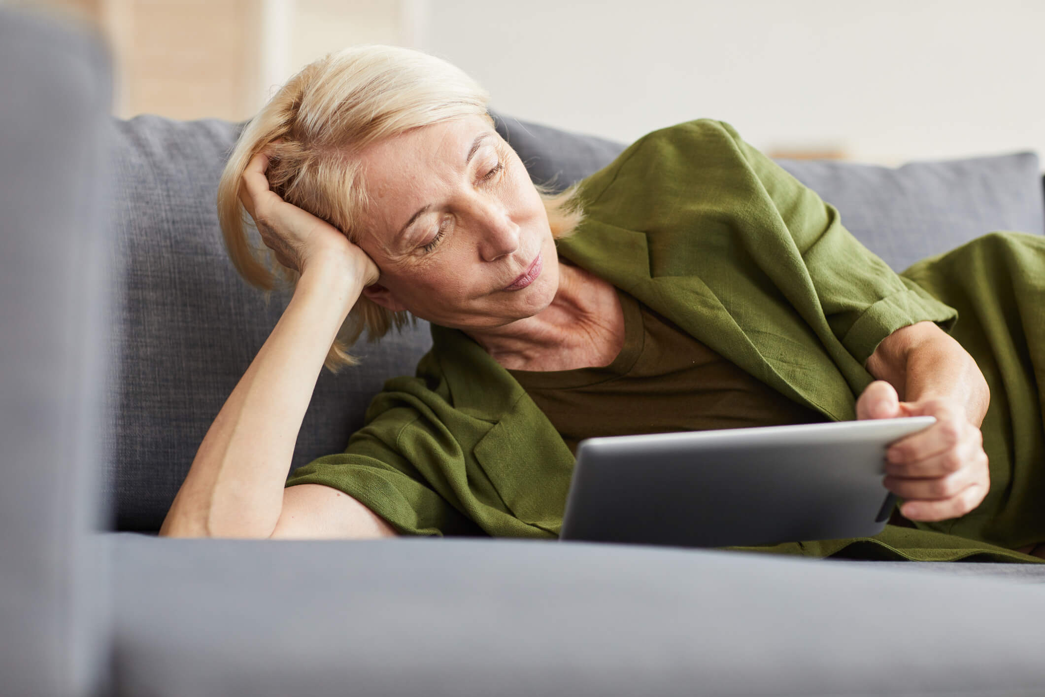 A woman is leaning on her arm on a couch and using a tablet; she has a relaxed expression.
