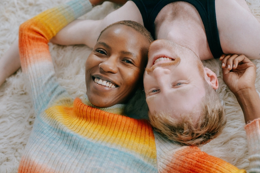 A couple are lying on the floor, with their heads together and their arms touching; they are both smiling.