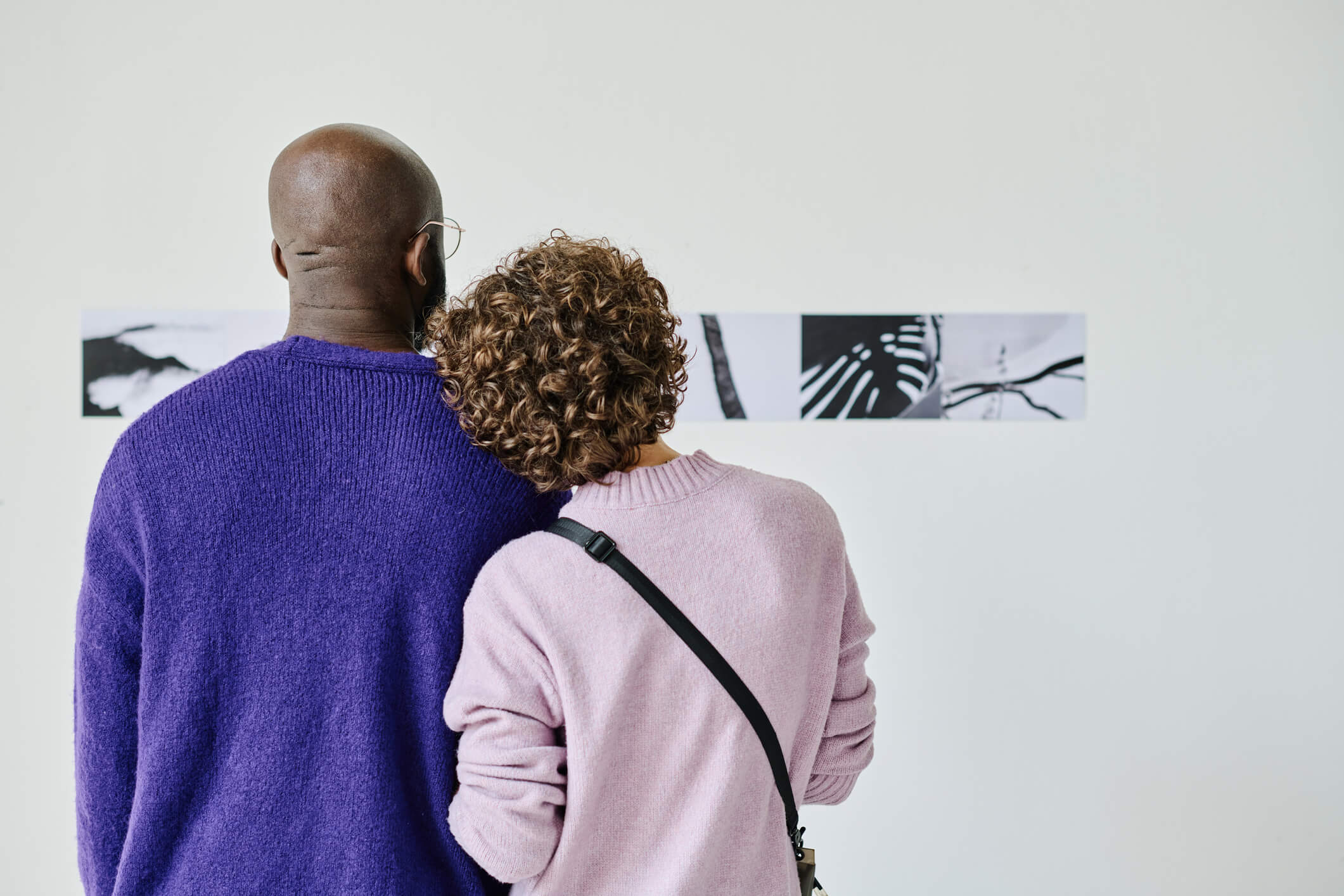 A couple wearing sweaters are standing up and looking at artwork on the wall in a museum; the woman has her head on the man’s shoulder.