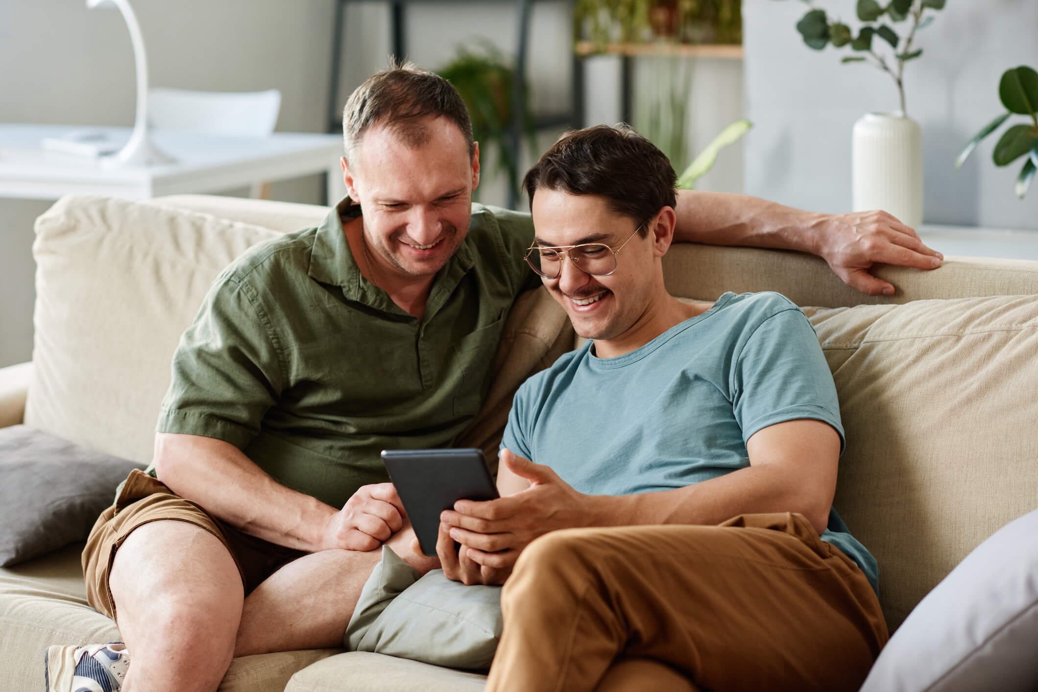 Two men are sitting on a couch and looking at a tablet screen; they are both smiling.