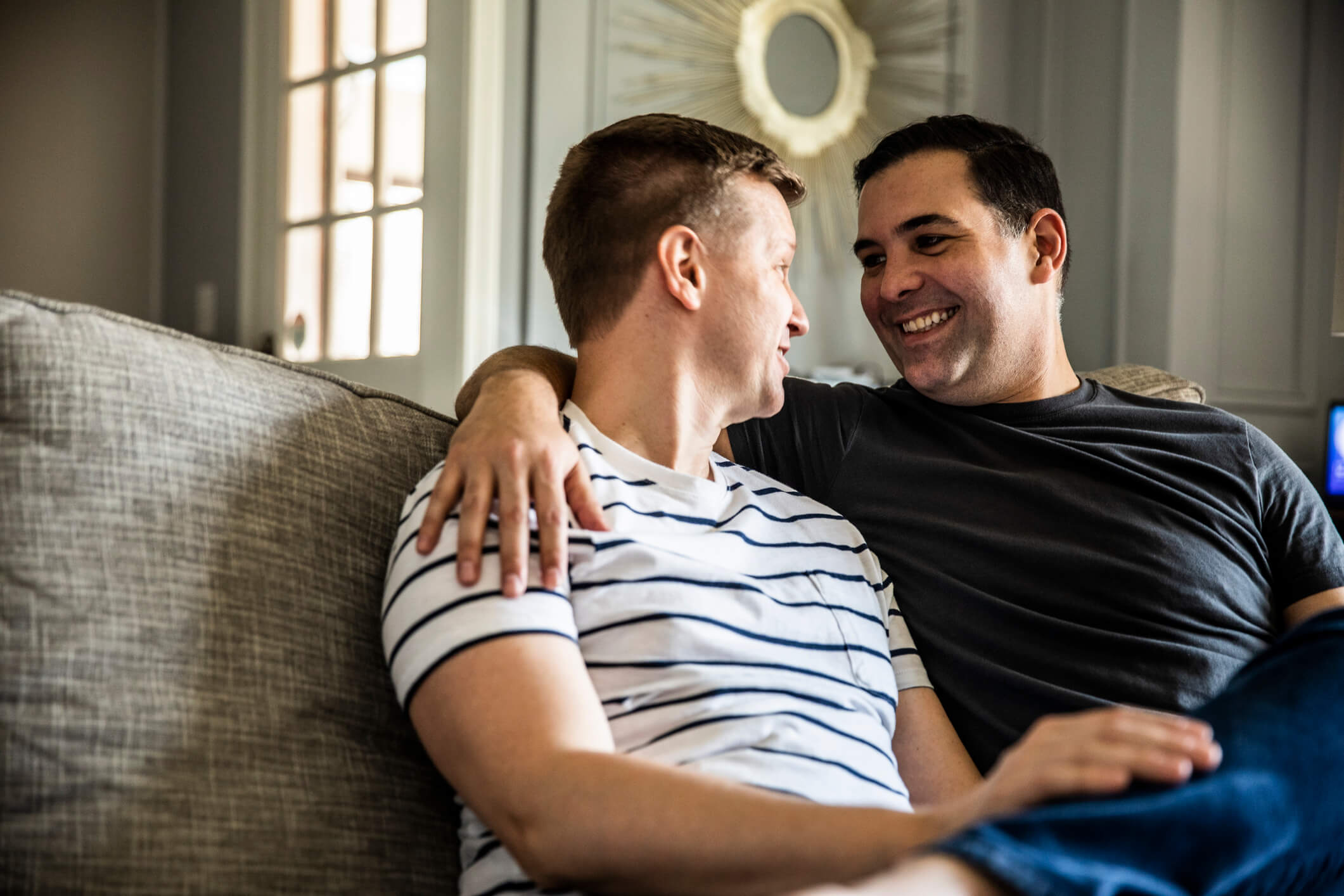 Two men are sitting on a couch and looking into each other’s eyes; one has his arm around the other one and they are both smiling.