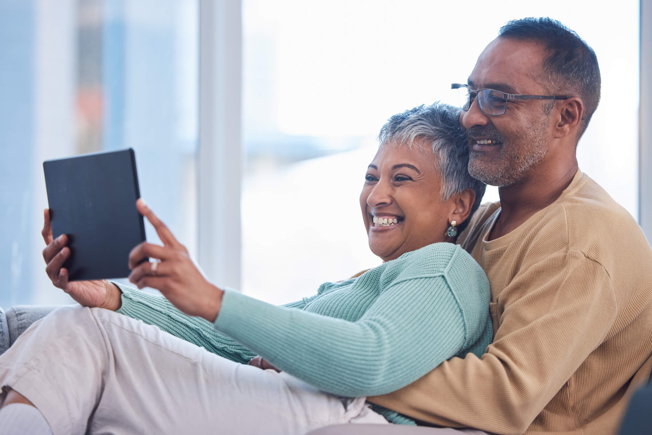 A couple are lying on a couch and looking at a tablet screen; the man is hugging the woman and the woman is holding onto the tablet, and they are both smiling.