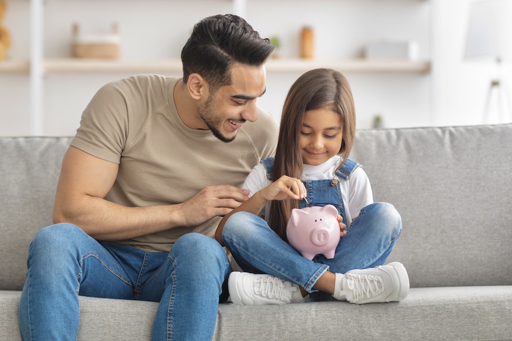 A father and his daughter are sitting on a couch; he is leaning over and has his hand on her shoulder, and she is putting money in a piggy bank, they are both smiling.