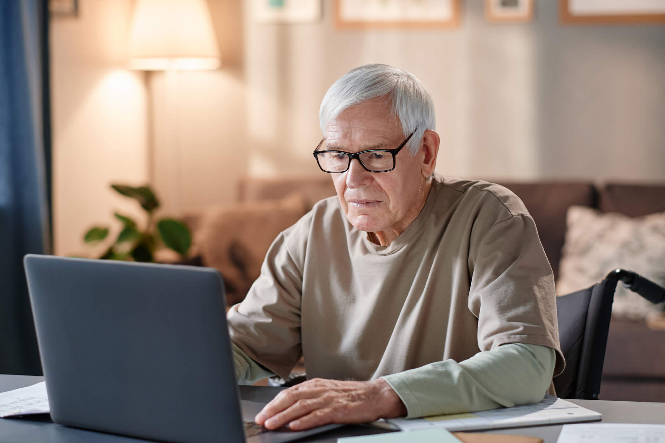 An older man in a wheelchair is at a table using a laptop and he has a pensive expression.