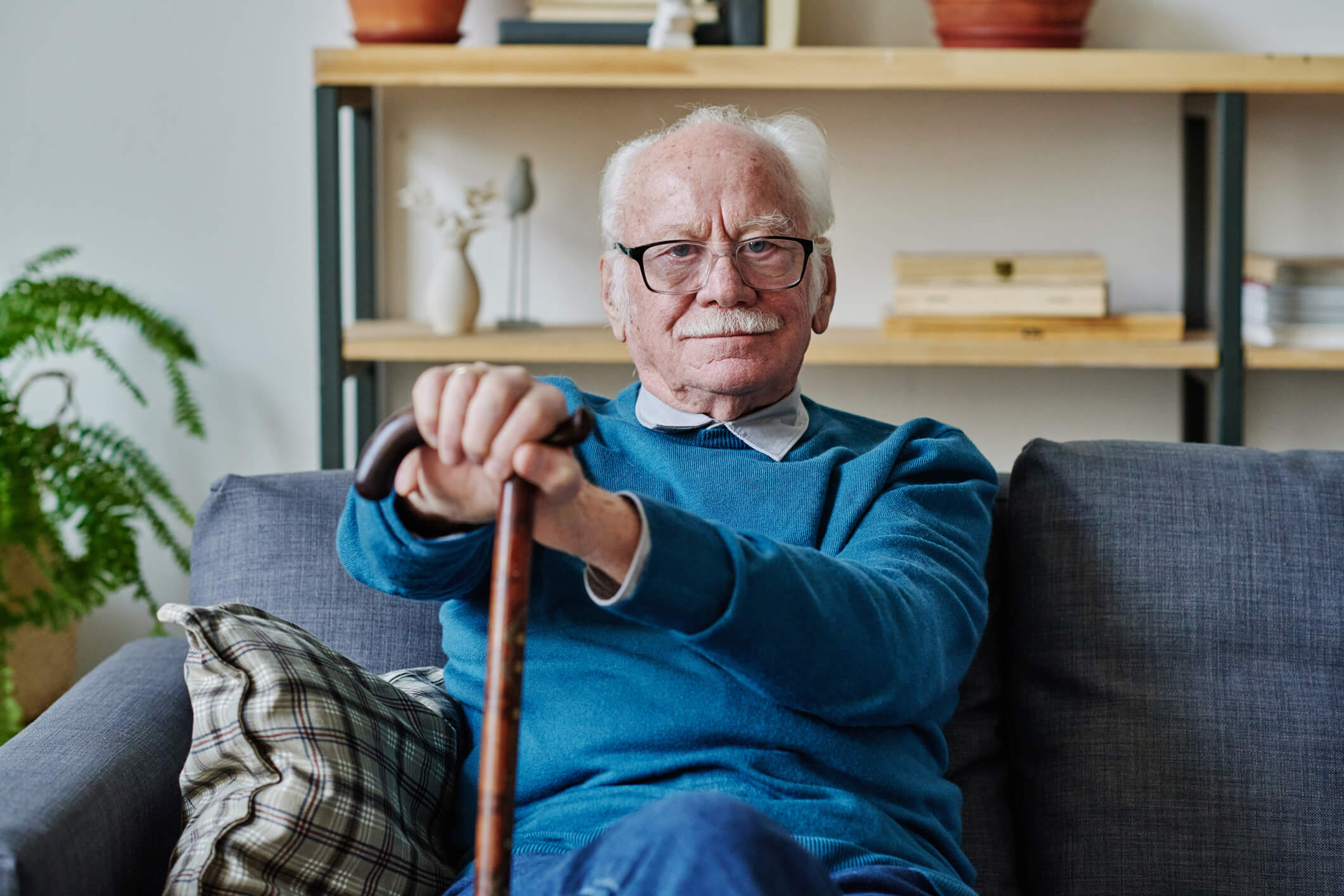 An older man is sitting on a couch and looking straight ahead; he is holding a cane and is smiling.