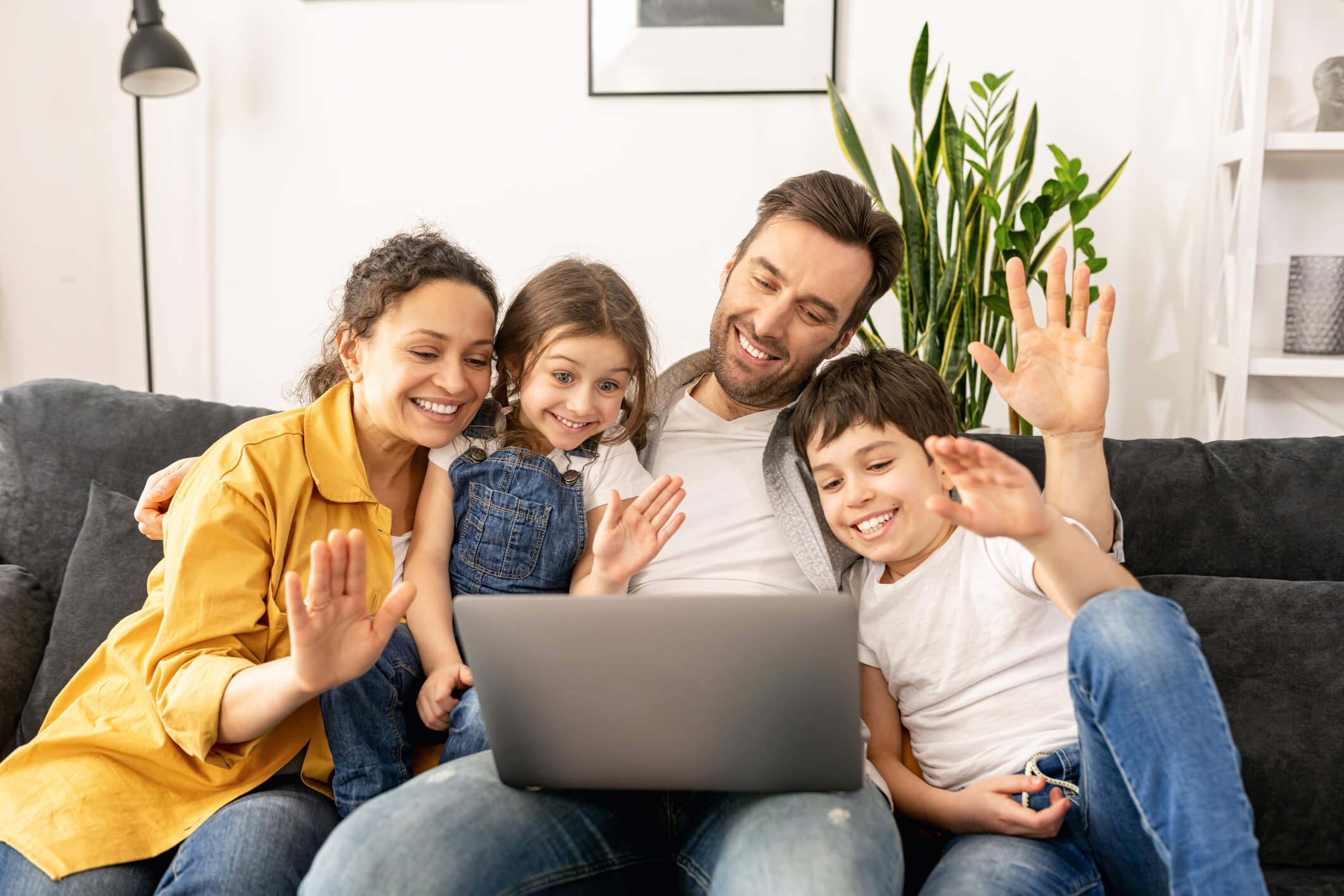 A family of four are sitting together on a couch and looking at a laptop screen; they are waving and smiling.