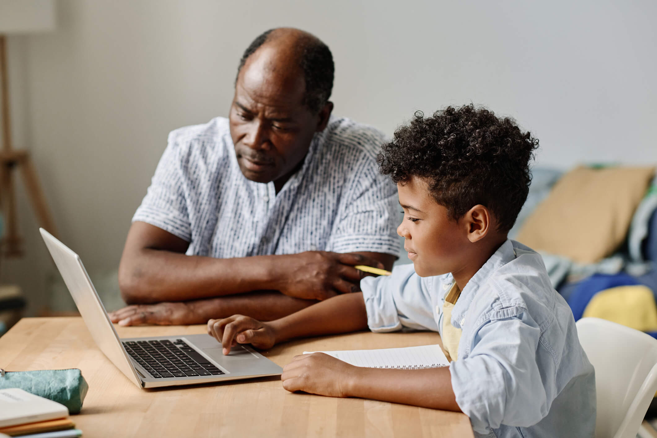 A dad and his son are sitting at a desk doing homework; the son is using a laptop, while dad supervises.