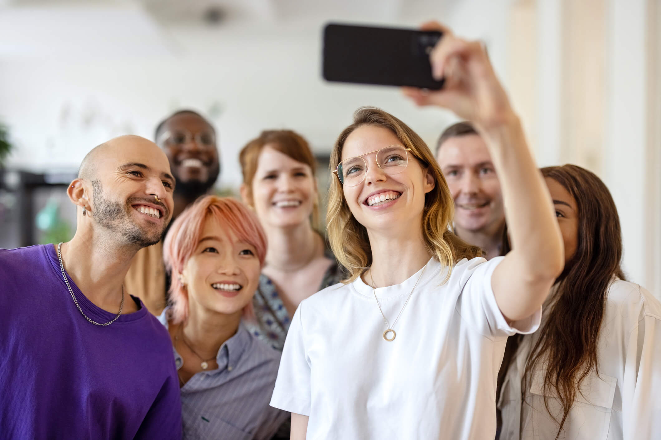 A group of friends are standing up in a room together and taking a selfie; one person is holding the phone up to take a picture, and everyone is smiling.