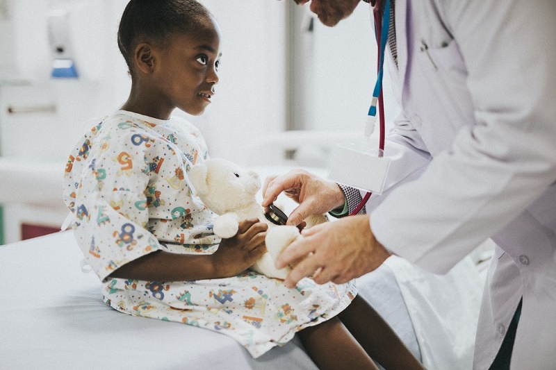 A child is sitting on a hospital bed with a teddy bear, and a TRICARE doctor is standing in front of her; she is looking up at the Tricare doctor.