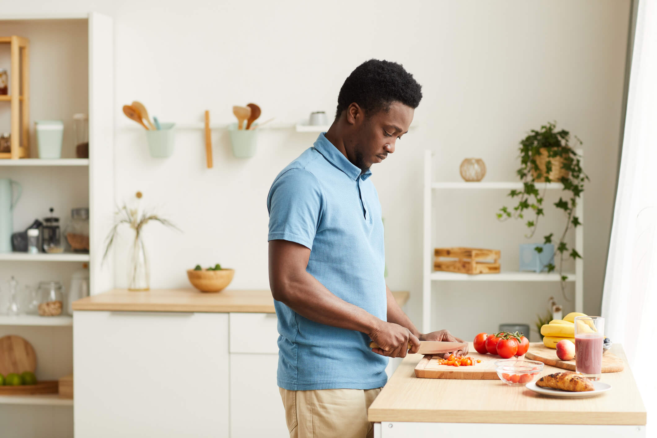 A man wearing a blue collared shirt is standing in front of the kitchen counter and chopping tomatoes with a focused expression.