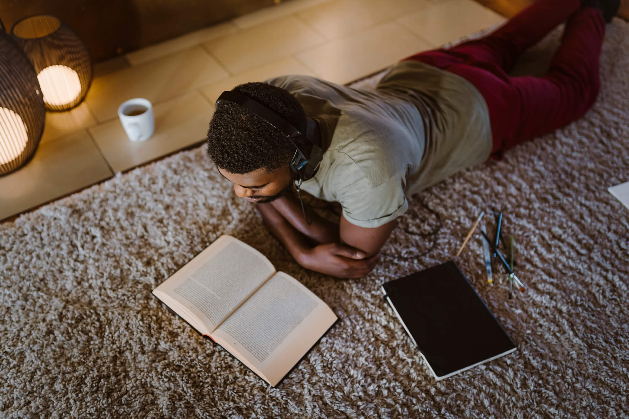 A man wearing headphones and a gray shirt lies on his stomach on a carpeted floor while reading a book on child development.