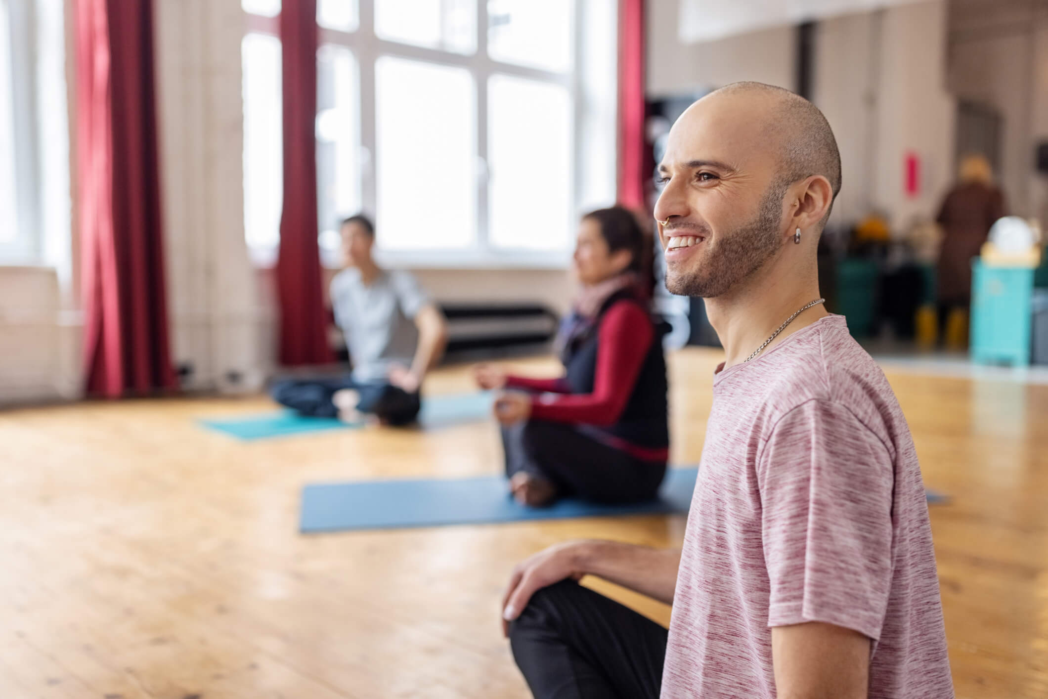 A man is sitting on a yoga mat in a class of other students sitting on yoga mats; he is looking to the side and smiling.