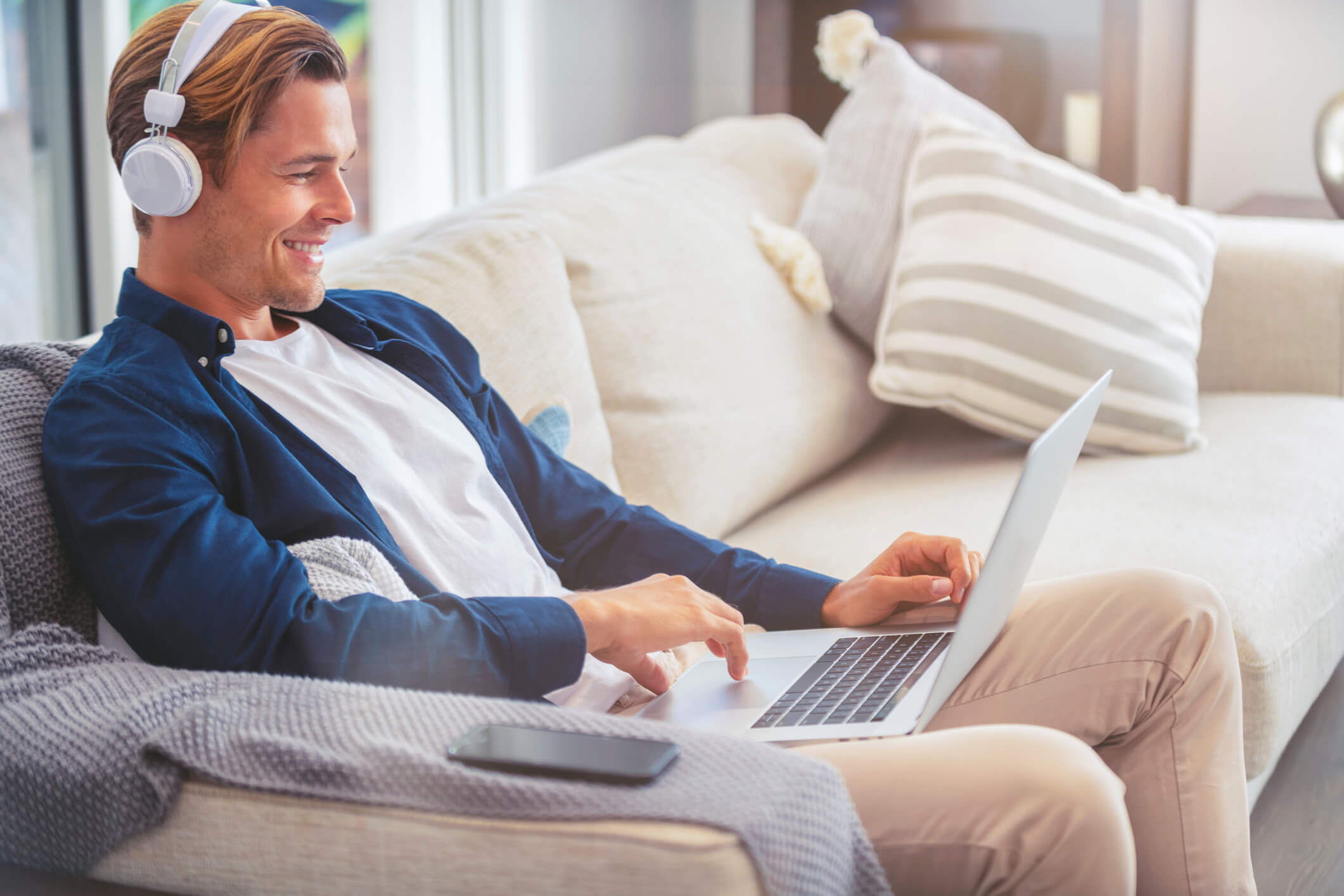 A man wearing headphones is sitting on a couch and using a laptop that is on his legs; he is smiling.