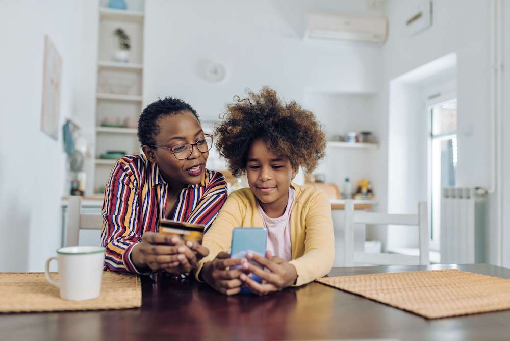 A mother and daughter are sitting at a table together; the mom is holding a credit card and the child is entering details into a phone.