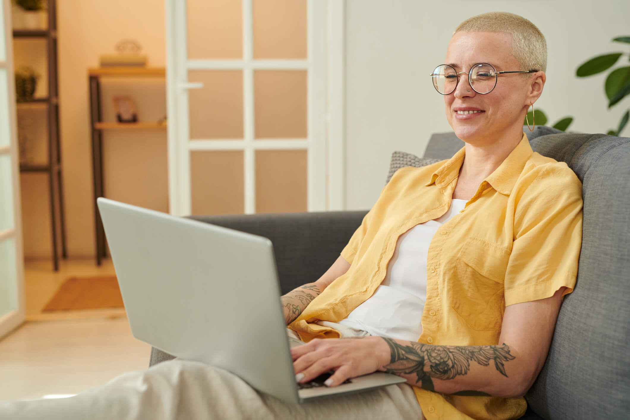 A person wearing glasses is sitting on a couch and using a laptop; they have a big smile on their face.