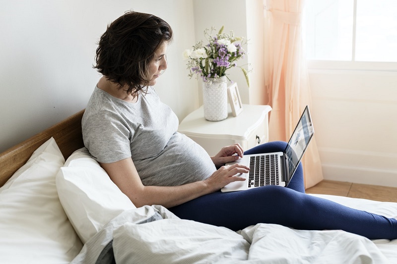 A pregnant woman is sitting up in bed and is using a laptop; she has an interested expression on her face.