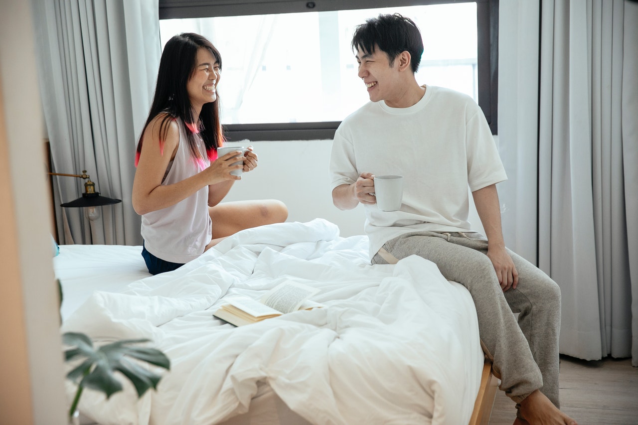 A couple are sitting on a bed with coffee cups in their hands; they both have big smiles on their faces.