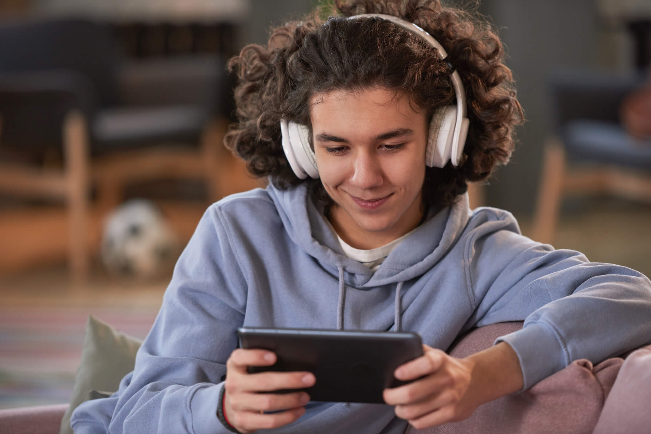 A teenager wearing headphones is sitting on a couch and using a tablet; he is also smiling.