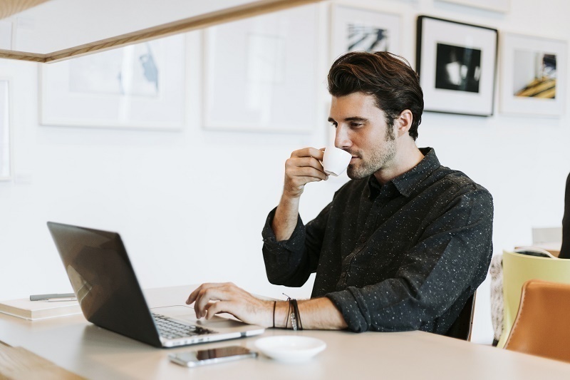 A man sits at his computer and sips an espresso in a coffee shop. He wears a black button down collared shirt.