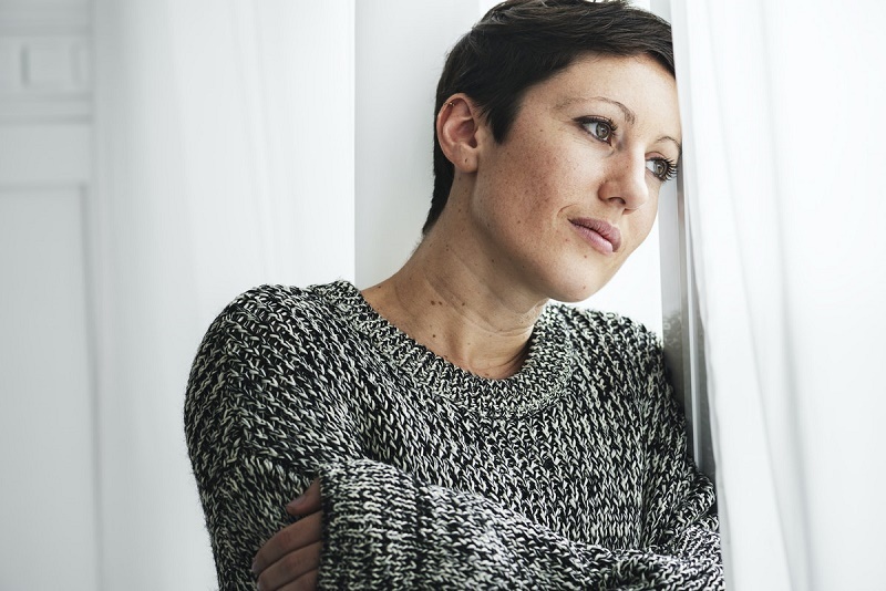 A woman in a sweater is standing up next to a window, and she has a forlorn expression on her face.