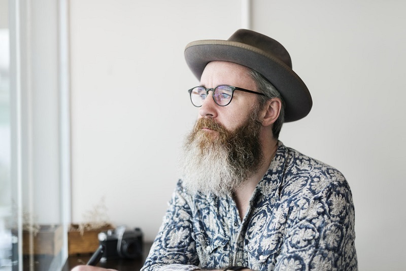 A man in a hat and collared shirt is looking out the window in a bright room and thinking about someone.