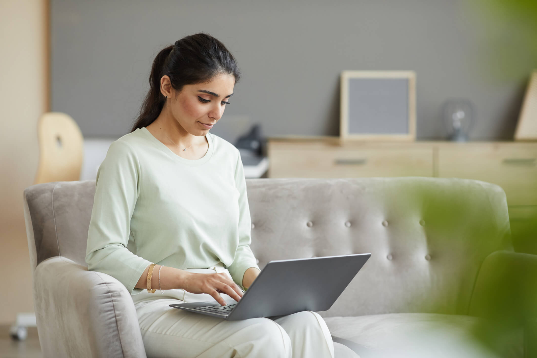 A woman with long hair and wearing a white long-sleeve blouse sits on the couch while searching for a mental health counselor on her computer.
