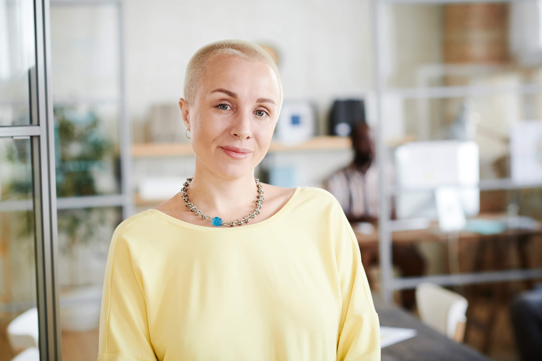 A person with short blonde hair, necklace, and yellow top smiles gently at the camera in a brightly lit office space.